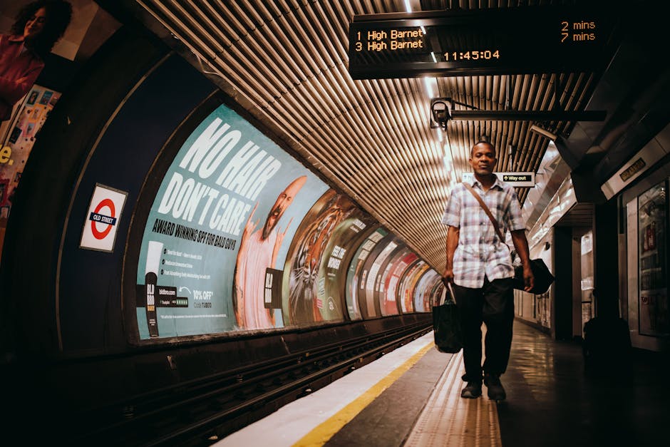 A man walking along a London Underground train station platform with a black briefcase in his right hand and a black shoulder bag across his body. He is wearing a short-sleeved checked shirt and dark