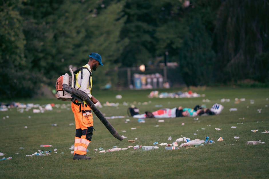 A person standing outdoors on a grassy area is wearing a yellow and gray checkered shirt over a green top, with green gloves on their hands. They are holding open a large black rubbish bag, which is p